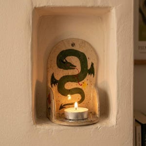 May include: A ceramic candle holder with a green dragon design. The dragon has a red tongue and black wings. A lit tea light candle sits in front of the dragon. The candle holder is set in a white alcove.