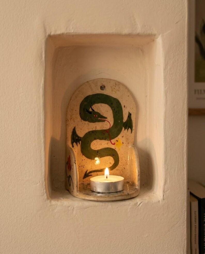 May include: A ceramic candle holder with a green dragon design. The dragon has a red tongue and black wings. A lit tea light candle sits in front of the dragon. The candle holder is set in a white alcove.