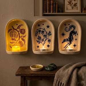 Może przedstawiać: Trzy kremowe ceramiczne kinkiety ścienne z łukowatymi górnymi częściami, każdy z unikalnym wzorem: granatem, wzorem kwiatowym i skaczącym psem. Każdy kinkiet zawiera zapaloną świecę, która daje ciepłą poświatę. Wzory są ciemnobrązowe i czarne.