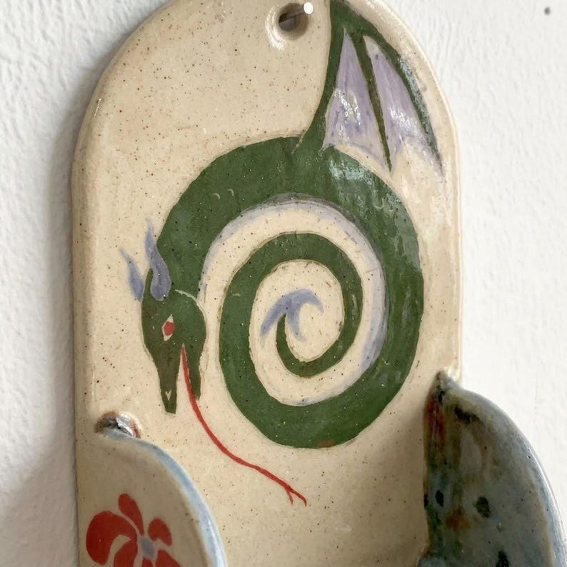May include: Ceramic wall hanging with a green dragon design. The dragon has a red tongue, blue horns, and purple wings. The background is a speckled beige color. The bottom has two small, curved holders, one with a red floral design.