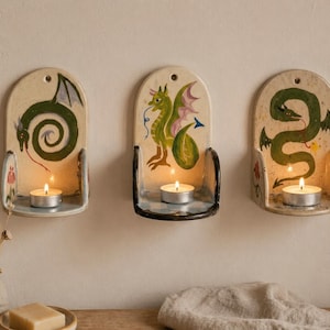 May include: Three ceramic wall sconces with dragon designs. Each sconce is off-white with a different green dragon illustration and a small metal candle holder. A lit tea light candle sits in each holder, casting a warm glow.