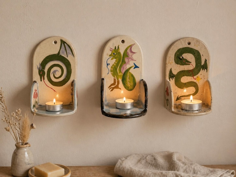 May include: Three ceramic wall sconces with dragon designs. Each sconce is off-white with a different green dragon illustration and a small metal candle holder. A lit tea light candle sits in each holder, casting a warm glow.