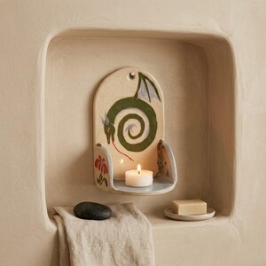 May include: A ceramic wall-mounted candle holder with a dragon design in green, red, and gray. The holder has a small shelf with a lit white candle. A red flower is painted on the side. The holder is mounted in a beige alcove.