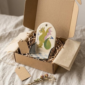 May include: A ceramic dragon-themed soap dish, hand-painted with green, purple, and blue details, is presented in a cardboard gift box with shredded paper. The dish features a dragon design and a checkered pattern. Includes a blank tag.