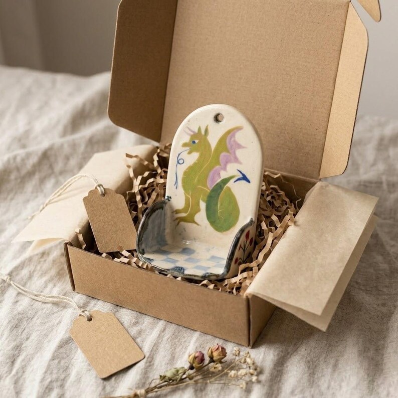 May include: A ceramic dragon-themed soap dish, hand-painted with green, purple, and blue details, is presented in a cardboard gift box with shredded paper. The dish features a dragon design and a checkered pattern. Includes a blank tag.