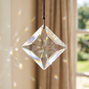 May include: A clear, faceted crystal prism, square-shaped, hangs from a brown cord. The crystal reflects light, creating a rainbow effect. The background includes a window and neutral-colored curtains, suggesting a decorative item for home decor.