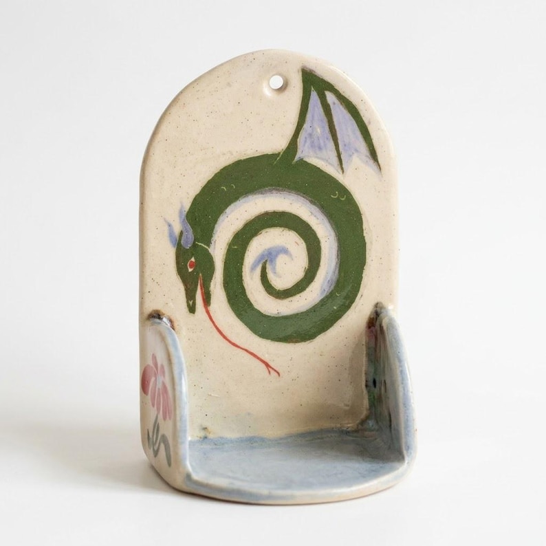 May include: A ceramic wall-mounted soap dish with a whimsical dragon design. The dragon is green with blue wings and a red tongue, set against a cream-colored background. The dish has a light blue base and a small hole for hanging.