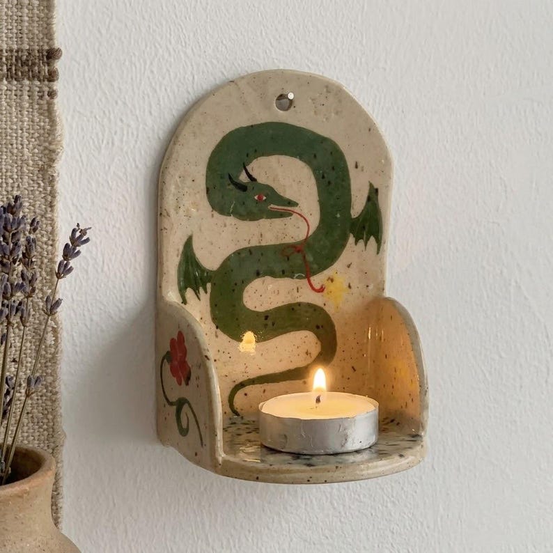 May include: A ceramic wall-mounted candle holder with a green dragon design. The dragon has a long, winding body and a red tongue. A small, lit candle sits in the holder, casting a warm glow. The holder is beige with a speckled texture.