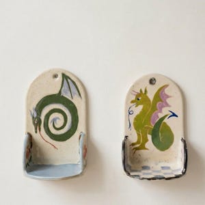 May include: Two ceramic wall-mounted spoon rests. One features a green dragon in a spiral, the other a green and yellow dragon with purple wings. Both have a small shelf and a hole for hanging.