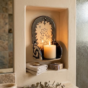 May include: A ceramic candle holder with a floral design and a lit candle. The holder is black and beige, with a shelf to hold the candle. Below, there are folded towels, a stone, and a bar of soap. The scene is set in a recessed wall niche.