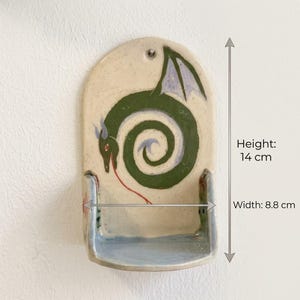 May include: Handmade ceramic wall pocket with a whimsical green dragon design. The pocket is 5.5 inches tall and 3.5 inches wide. The dragon has purple wings and a red tongue. The pocket has a light beige background and a blue base.