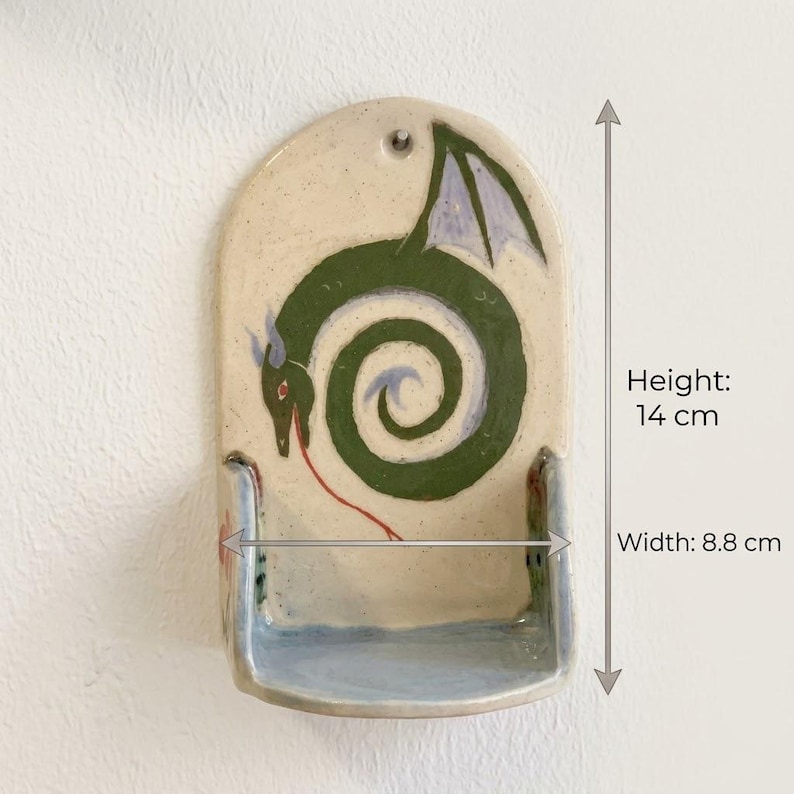 May include: Handmade ceramic wall pocket with a whimsical green dragon design. The pocket is 5.5 inches tall and 3.5 inches wide. The dragon has purple wings and a red tongue. The pocket has a light beige background and a blue base.
