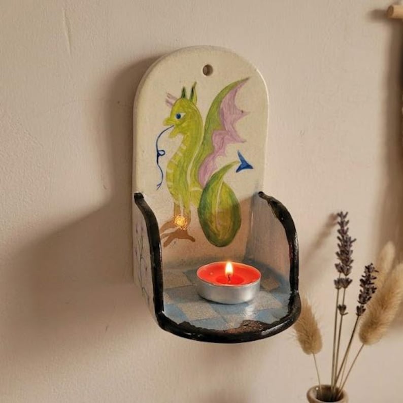 May include: A ceramic wall-mounted candle holder with a hand-painted dragon design. The dragon is green with pink wings. The holder has a black rim and a blue and white checkered base, with a lit red tea light candle inside.