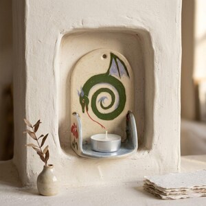May include: A ceramic candle holder with a green dragon design. The dragon is painted on a cream-colored background, with a red tongue and blue wings. A small tea light candle sits on a shelf below the dragon. The holder is set into a white wall.