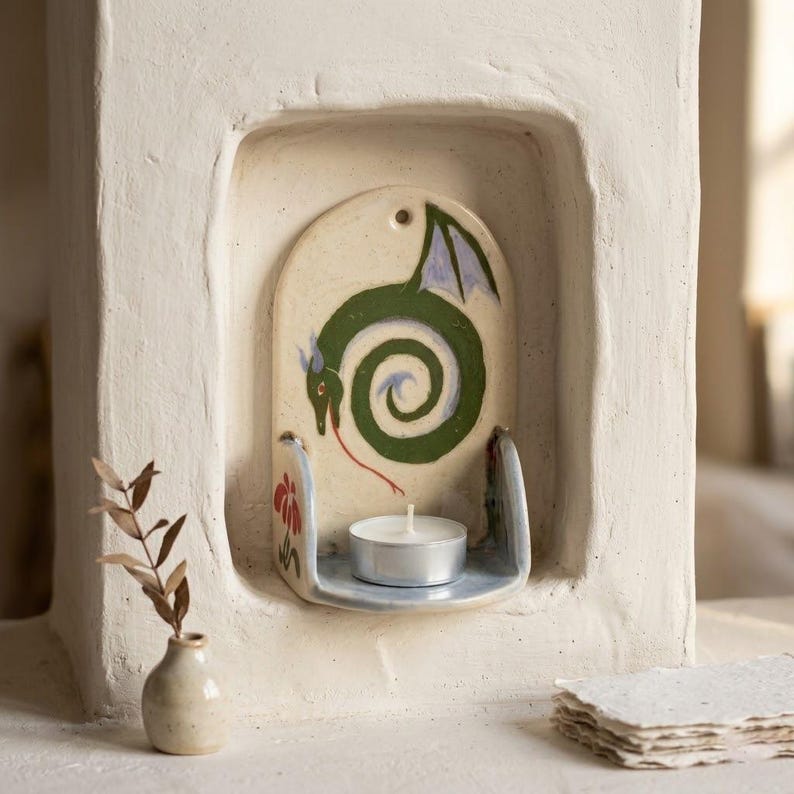 May include: A ceramic candle holder with a green dragon design. The dragon is painted on a cream-colored background, with a red tongue and blue wings. A small tea light candle sits on a shelf below the dragon. The holder is set into a white wall.