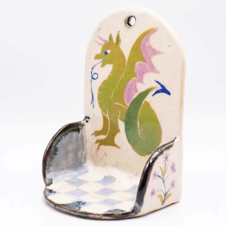 May include: Ceramic wall pocket with a whimsical dragon design. The dragon is green with pink wings and blue accents. The pocket has a checkered base and floral details, with a hole for hanging.