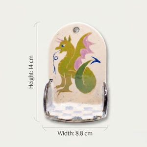 May include: A ceramic wall pocket, 5.5 inches tall and 3.5 inches wide, featuring a whimsical dragon design. The dragon is green with pink wings and a blue tongue. The pocket has a checkered base and a hole for hanging.