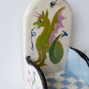 May include: A ceramic wall pocket with a whimsical dragon design. The dragon is green with purple wings and blue eyes. The pocket has a light blue and white checkered pattern on the bottom and a floral design on the side.