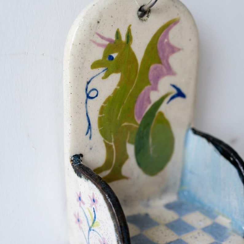 May include: A ceramic wall pocket with a whimsical dragon design. The dragon is green with purple wings and blue eyes. The pocket has a light blue and white checkered pattern on the bottom and a floral design on the side.