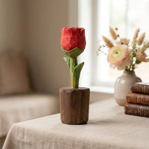 Può includere: Una scultura di rosa in legno intagliata a mano con un fiore rosso vibrante, foglie verdi e una base in legno marrone. La rosa è esposta su un tavolo con una tovaglia di colore neutro. La scultura è un oggetto decorativo.