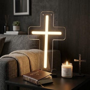 May include: A glowing cross-shaped LED lamp illuminates a dark room, next to a lit candle and a Bible. A small metal cross and a framed cross artwork are also visible. The lamp is on a black table.