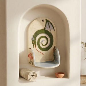 May include: A ceramic wall shelf with a dragon design. The shelf is cream-colored with a green dragon in a spiral pattern. A rolled towel and small bowl are in the alcove. A vase with greenery sits on a wooden shelf.