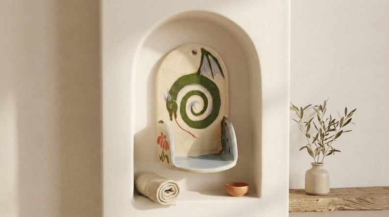 May include: A ceramic wall shelf with a dragon design. The shelf is cream-colored with a green dragon in a spiral pattern. A rolled towel and small bowl are in the alcove. A vase with greenery sits on a wooden shelf.