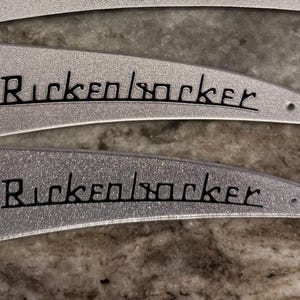 May include: Three silver-colored, triangular metal truss rod covers for a guitar, each featuring a textured finish and the black text Rickenbacker printed horizontally across the center, with small pre-drilled holes at the corners for mounting.