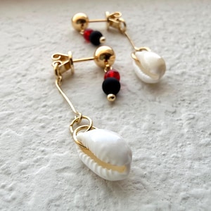 May include: A pair of gold-tone drop earrings featuring a white cowrie shell charm suspended from a thin metal wire. Each earring includes a small gold ball stud, a vibrant red bead, and a matte black bead positioned above the dangling shell charm.