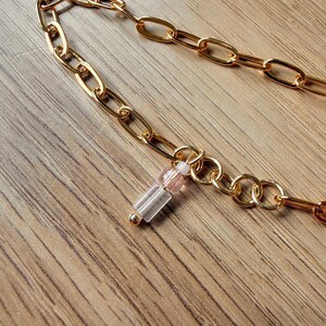 Pode incluir: Colar de corrente dourada com elos ovais. Um pequeno pingente com contas transparentes e rosa pende da corrente. O colar é exibido em uma superfície de madeira.