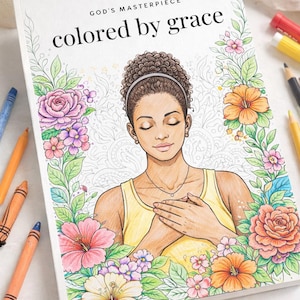 May include: A coloring book titled "colored by grace" with a floral border and an illustration of a woman with closed eyes. Crayons and colored pencils surround the book, suggesting a creative activity. The book's cover features the text "God's Masterpiece".