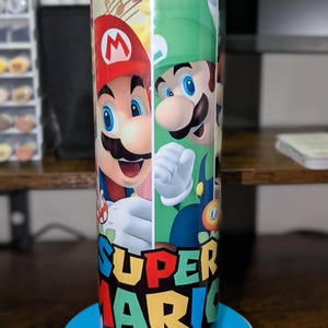 May include: A tall, cylindrical tumbler featuring a vibrant design of Mario and Luigi from Super Mario. The tumbler has a blue base and the words "SUPER MARIO" in colorful block letters. The characters are depicted in their classic outfits.