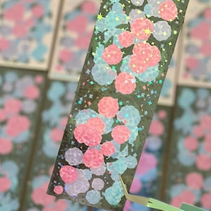 May include: A rectangular sticker with a holographic finish, featuring a pattern of pink and blue circular shapes and star-shaped accents. The sticker is held by green tweezers, with other similar stickers visible in the background.