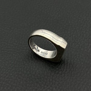 Könnte beinhalten: Ein silberner Ring mit einem einzigartigen, geformten Design. Der Ring hat eine flache, rechteckige Oberfläche und ein abgerundetes Band. Die Innenseite des Bandes ist mit Text und der Zahl 2016 graviert. Der Ring ist vor einem dunklen Hintergrund platziert.