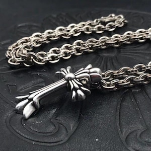 May include: A silver-colored cross pendant necklace with a detailed floral design. The cross is attached to a silver chain with a series of oval links. The necklace is displayed on a black surface.