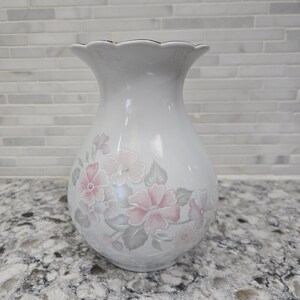 Peut inclure: Vase en céramique blanche avec un bord festonné et un motif floral. Le vase présente des fleurs roses et des feuilles grises. Le bord a une bordure dorée. Le vase mesure environ 20 cm de haut.