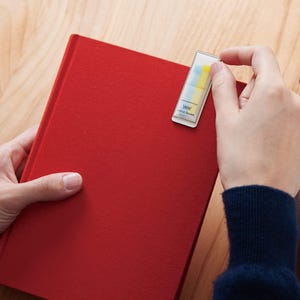 Puede incluir: Un cuaderno de tapa dura de color rojo brillante con un dispensador rectangular de pestañas adhesivas de plástico transparente en la esquina superior derecha. El dispensador contiene pequeñas banderas adhesivas azules y amarillas y muestra el texto coco fusen.
