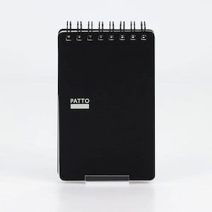 May include: A black spiral-bound notebook with a rectangular shape. The front cover features the word PATTO printed in white capital letters above a solid white horizontal rectangle. The notebook is held upright by a clear acrylic stand.