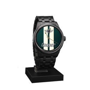 May include: Black wristwatch with a round face featuring a green and white striped design and the word "Cooper." The watch has a black metal band and is displayed on a black stand. The watch face includes the Mini logo.