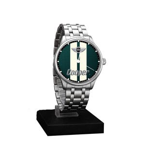 May include: A silver wristwatch with a green and white face, featuring the word "Cooper" and the Mini logo. The watch has a stainless steel band and is displayed on a black stand.