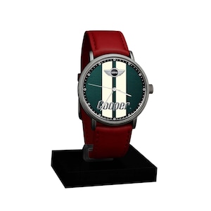 May include: A wristwatch with a red leather band and a silver-colored case. The watch face features a green background with white stripes and the word "Cooper" in white. The watch is displayed on a black stand.
