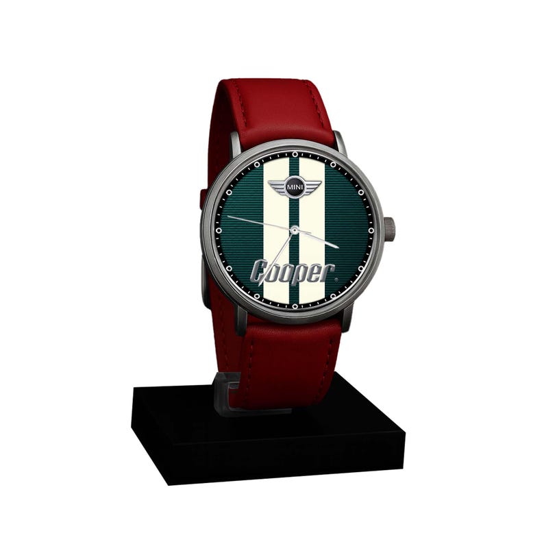 May include: A wristwatch with a red leather band and a silver-colored case. The watch face features a green background with white stripes and the word "Cooper" in white. The watch is displayed on a black stand.