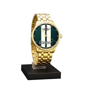 May include: A gold-toned wristwatch with a green and white face, featuring the "MINI Cooper" logo. The watch has a gold band and is displayed on a black stand. The watch face has a green background with two white vertical stripes and the word "Cooper".