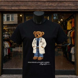 May include: Black t-shirt featuring a brown bear wearing a white lab coat, blue jeans, and a stethoscope. The text "POLO BEAR by RALPH LAUREN" and signature are printed at the bottom. The store name "URWAH ALBARIQI STORE" is at the top.