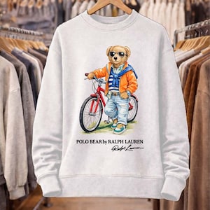 May include: White sweatshirt featuring a cartoon bear wearing sunglasses, an orange jacket, and blue jeans, riding a red bicycle. The text "POLO BEAR by RALPH LAUREN" and signature are printed below the image. The sweatshirt is hanging on a wooden hanger.