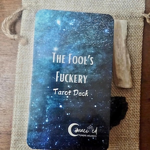 Op de afbeelding: Een rechthoekig tarotkaartendeck met een donkerblauwe sterrenstelselprint en witte sterren. De tekst luidt The Fool's Fuckery Tarot Deck en Connected Fungirl Wellness. Het deck ligt op een beige jute zakje met een licht houten stokje en een zwarte steen.