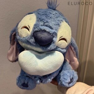 May include: A plush blue Stitch doll with large white eyes, a dark blue nose, and a light blue heart-shaped mouth. The doll has large ears and a tuft of hair on top of its head. The brand name "ELUROCO" is visible in the top right corner.