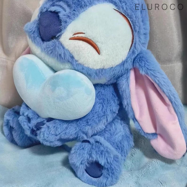 May include: A plush blue Stitch doll holding a light blue heart. The doll has large ears with pink insides, a white face, and brown stitched details. The word "ELUROCO" is visible in the top right corner.