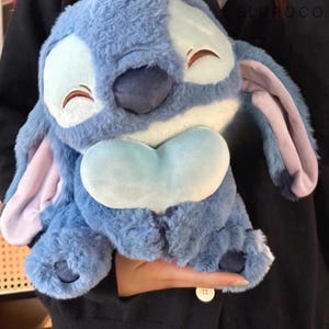 May include: A plush blue Stitch character with large ears, a heart-shaped belly, and a smiling expression. The plush toy has a soft, furry texture and is a popular character from a well-known animated film.