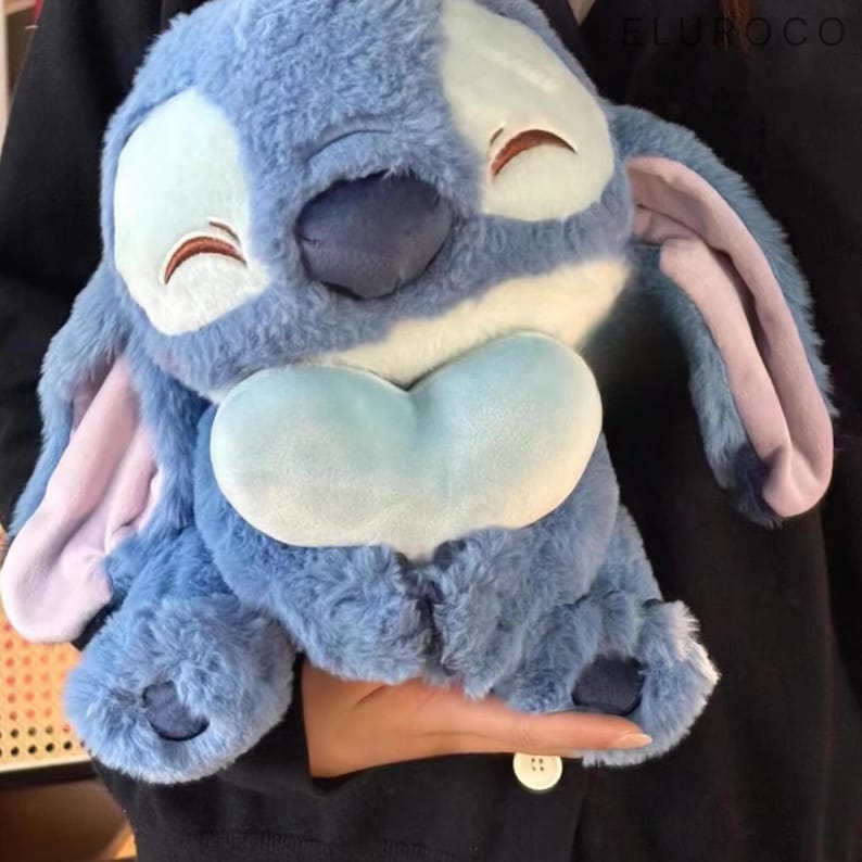 May include: A plush blue Stitch character with large ears, a heart-shaped belly, and a smiling expression. The plush toy has a soft, furry texture and is a popular character from a well-known animated film.